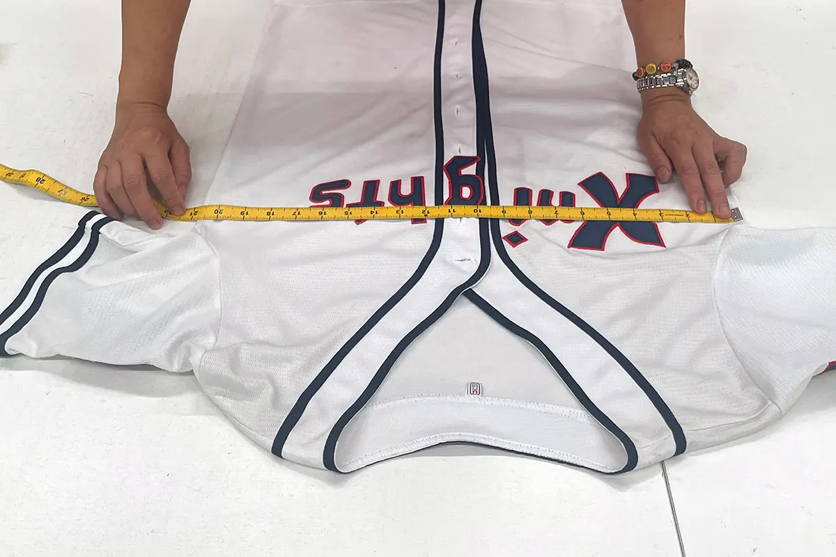 Hands measuring the chest width of a white baseball jersey laid flat